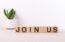 Join us sign on wooden cubes on a light table with flower and light background.