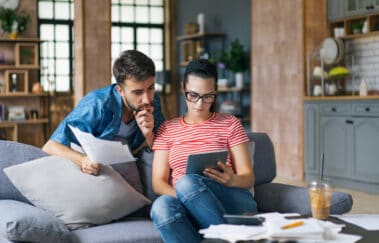 Couple calculating bills at home using tablet and calculator. Young couple working on computer while calculating finances sitting on couch. Young man with wife at home analyzing their finance with documents.