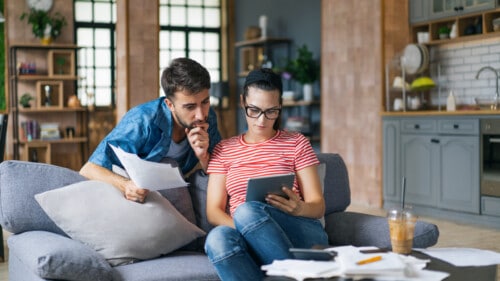 Couple calculating bills at home using tablet and calculator. Young couple working on computer while calculating finances sitting on couch. Young man with wife at home analyzing their finance with documents.